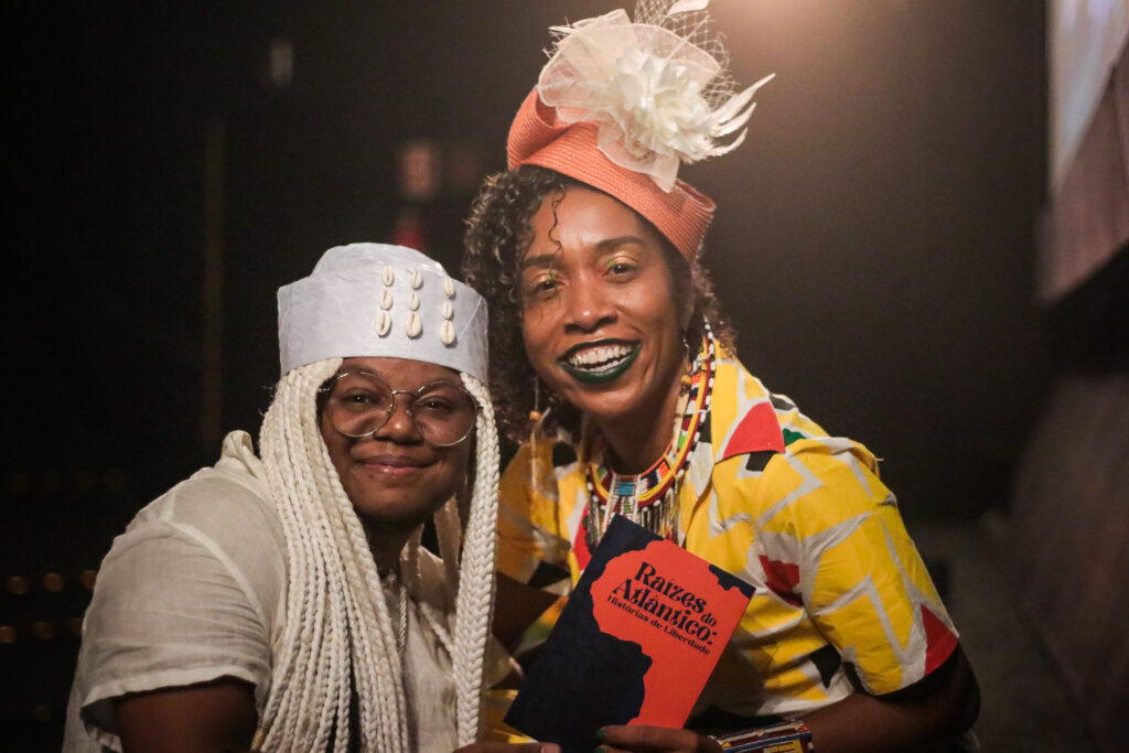 Homem negro trans usando roupa branca e uma mulher negra usando roupa colorida e um chapeu na cabeça laranja com branco